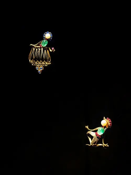 Two pins encrusted with diamonds and green, yellow, red and blue gemstones, depicting a scene of a woman on a balcony and a lover below